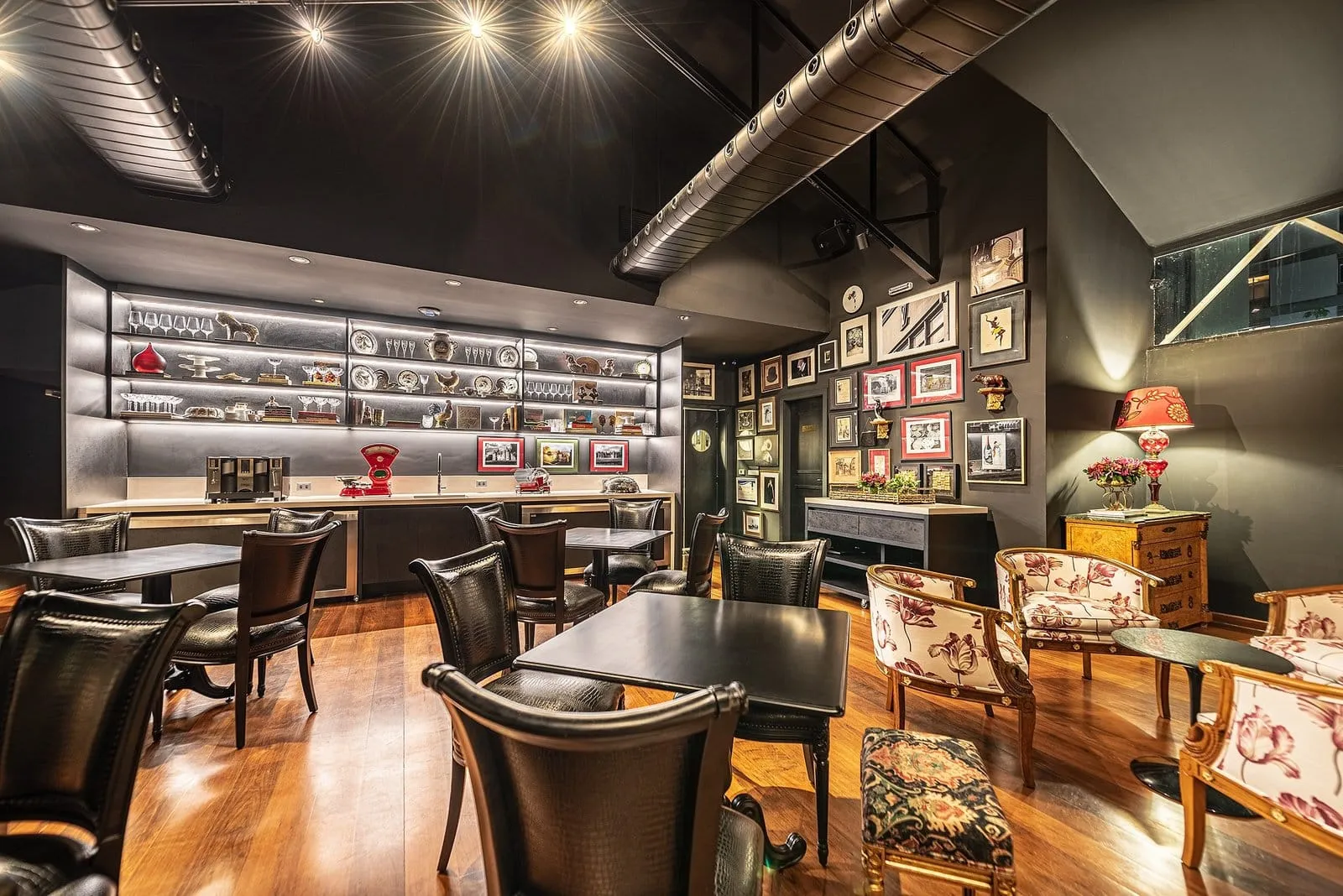 Salão de restaurante com estante iluminada, cadeiras de couro, quadros na parede e piso de madeira