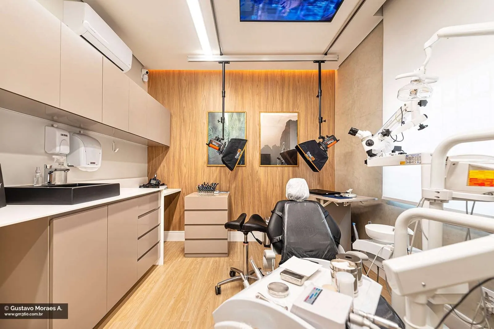Consultório com cadeira odontológica, armários brancos e pia sob janela iluminada
