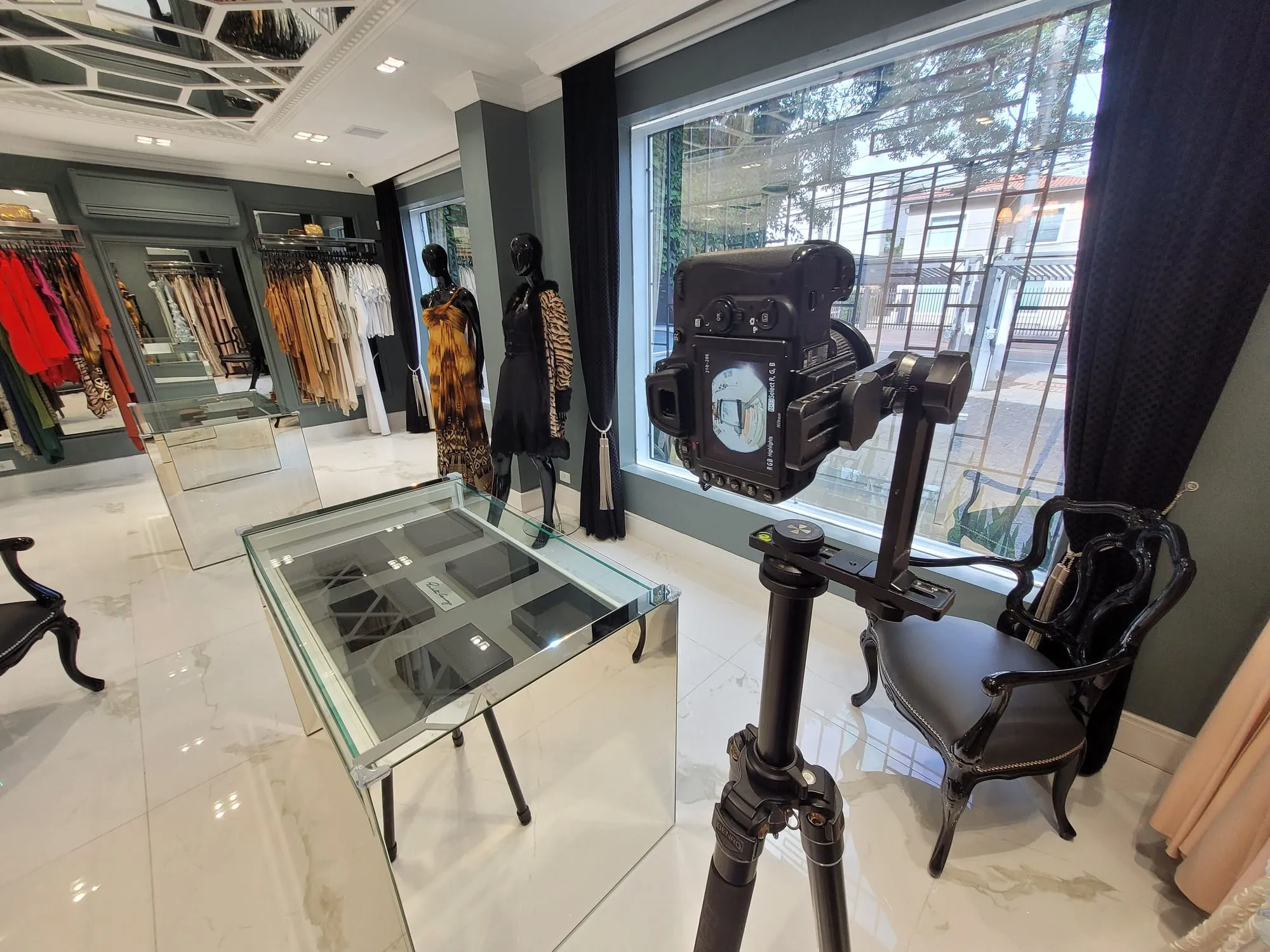 Câmera no tripé Benro junto à janela de grade decorativa do ateliê, ao lado de manequins vestidos, araras de roupas e uma vitrine de vidro