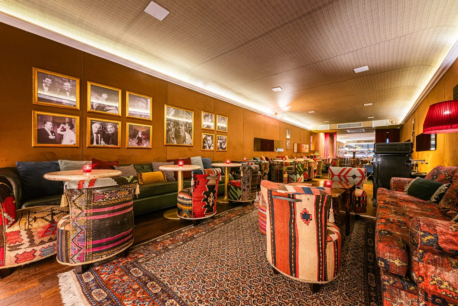 Poltronas forradas de kilim, mesas redondas de mármore com velas vermelhas e tapete persa, com parede de fotos emolduradas