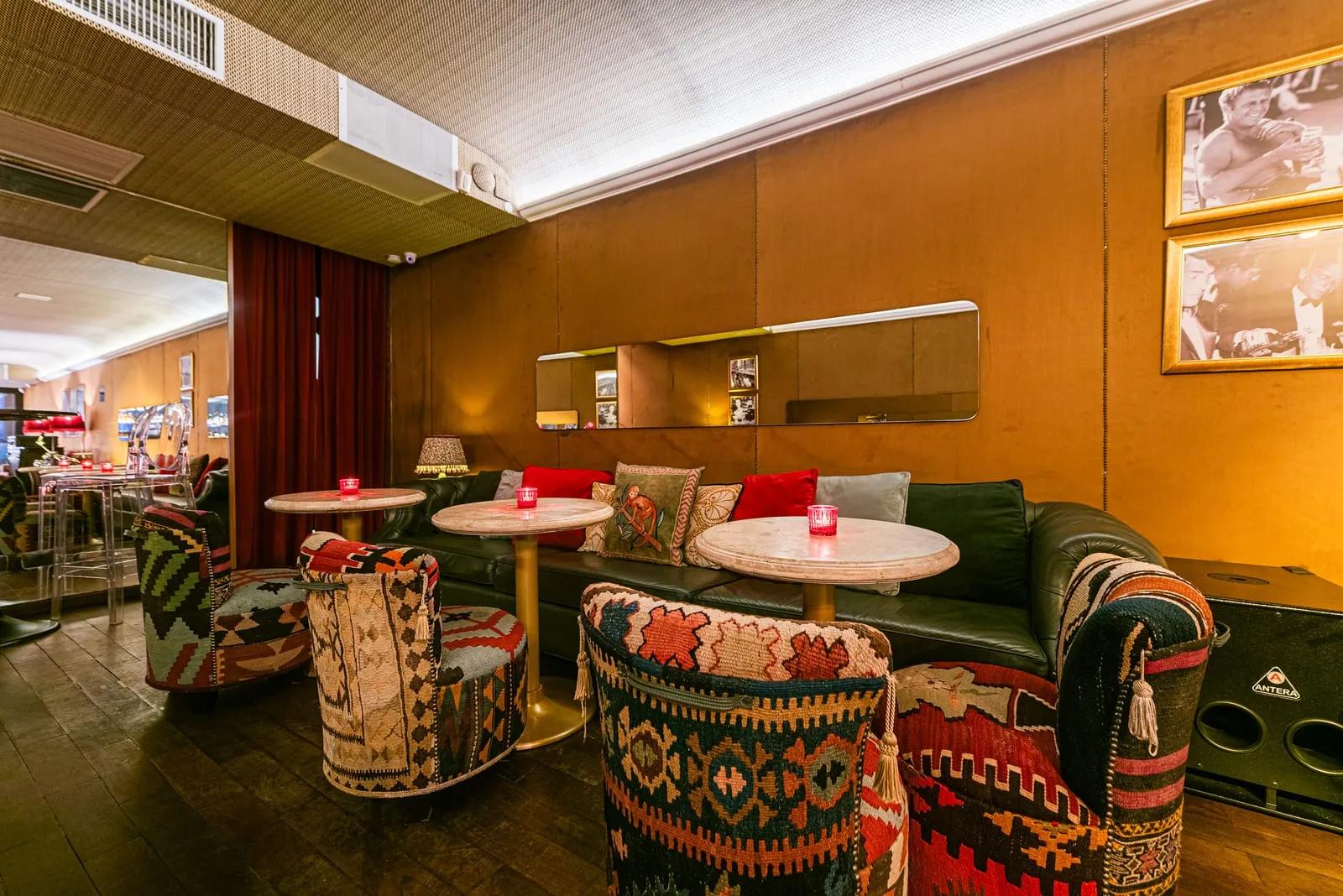 Sofá chesterfield de couro verde, poltronas de kilim e mesas de mármore, com cortina vermelha e espelho comprido na parede de madeira