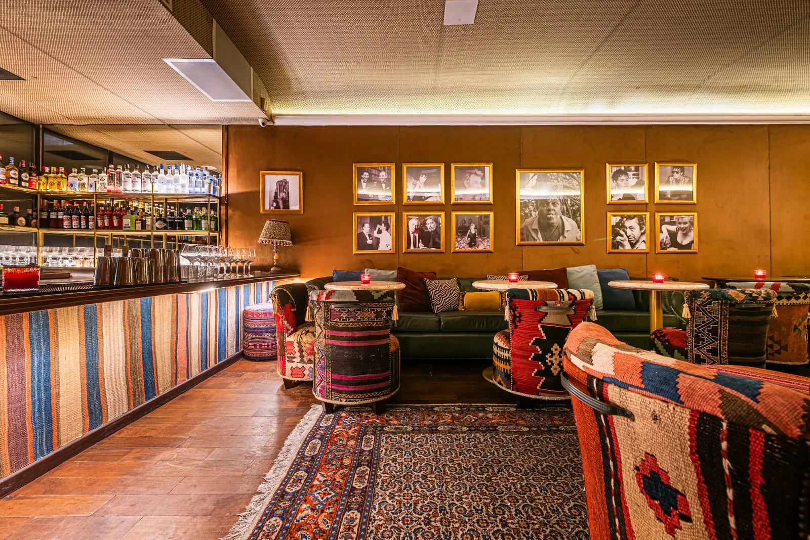 Balcão do bar revestido de tecido listrado, prateleiras de garrafas e taças, poltronas de kilim sobre tapete persa e parede de fotos