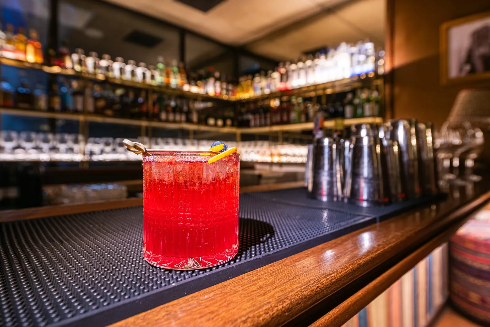 Drinque vermelho com gelo em copo lapidado sobre o balcão, com coqueteleiras e prateleiras de garrafas desfocadas ao fundo
