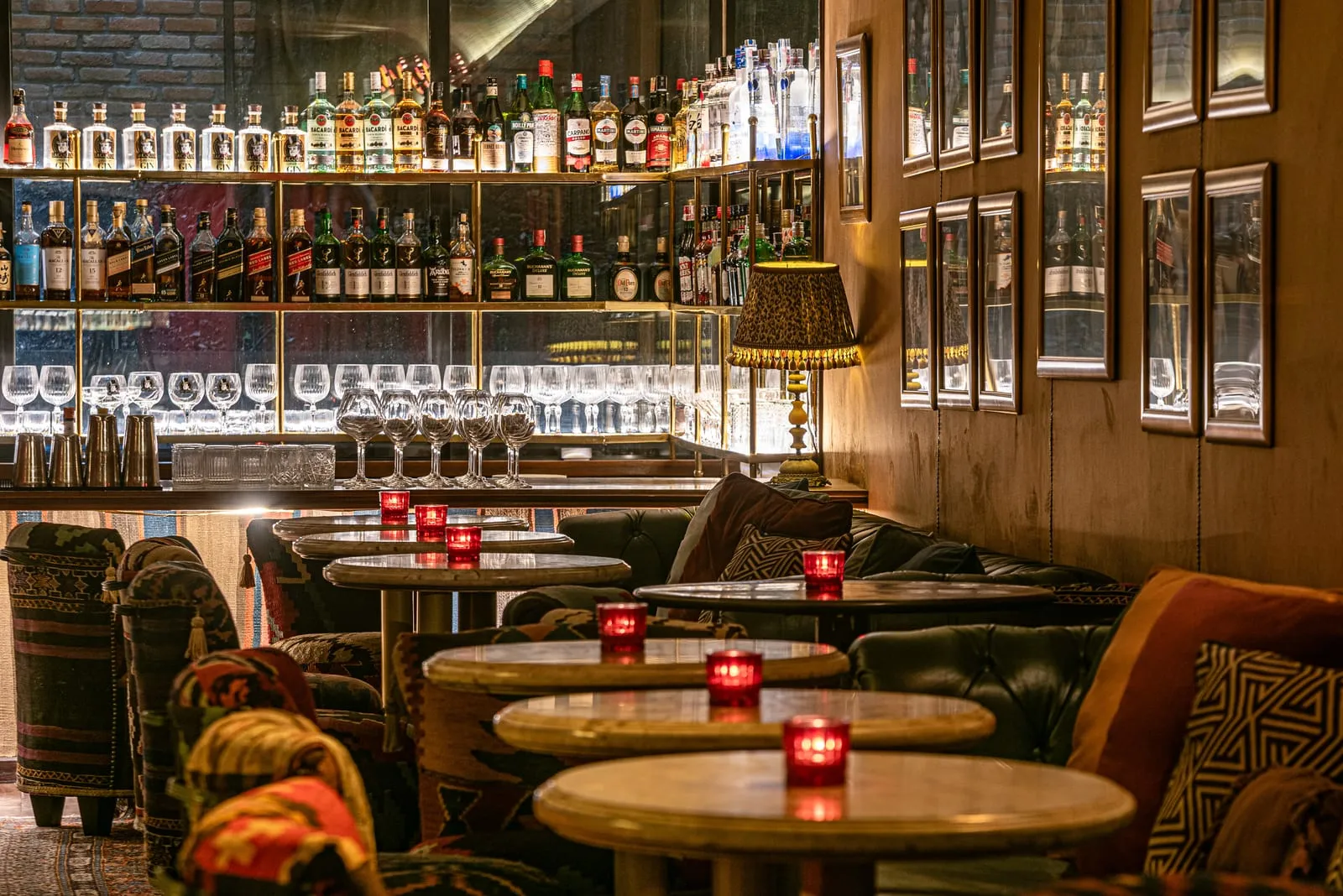 Fileira de mesas redondas com velas vermelhas diante das prateleiras de garrafas e taças iluminadas do bar