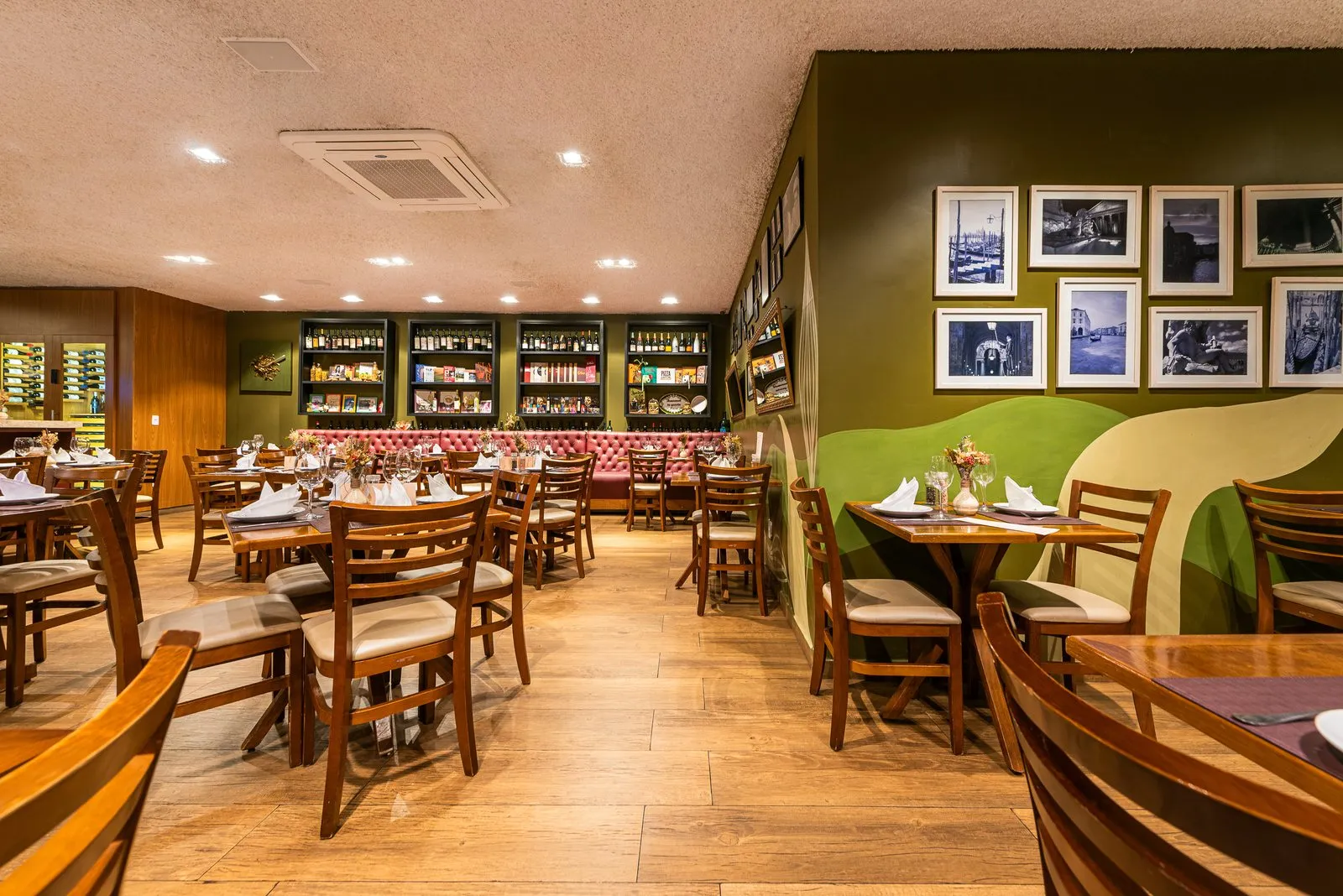 Salão da Cantina Castellammare com mesas de madeira, banco capitonê rosa sob estantes de vinhos e parede verde com fotos em preto e branco