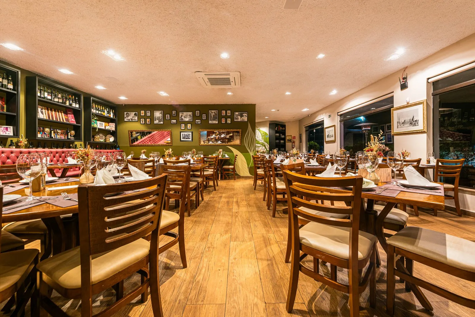 Salão da Cantina Castellammare com mesas de madeira postas, janelas de vidro à direita, paredes verde-oliva com fotos em preto e branco e estantes de vinhos
