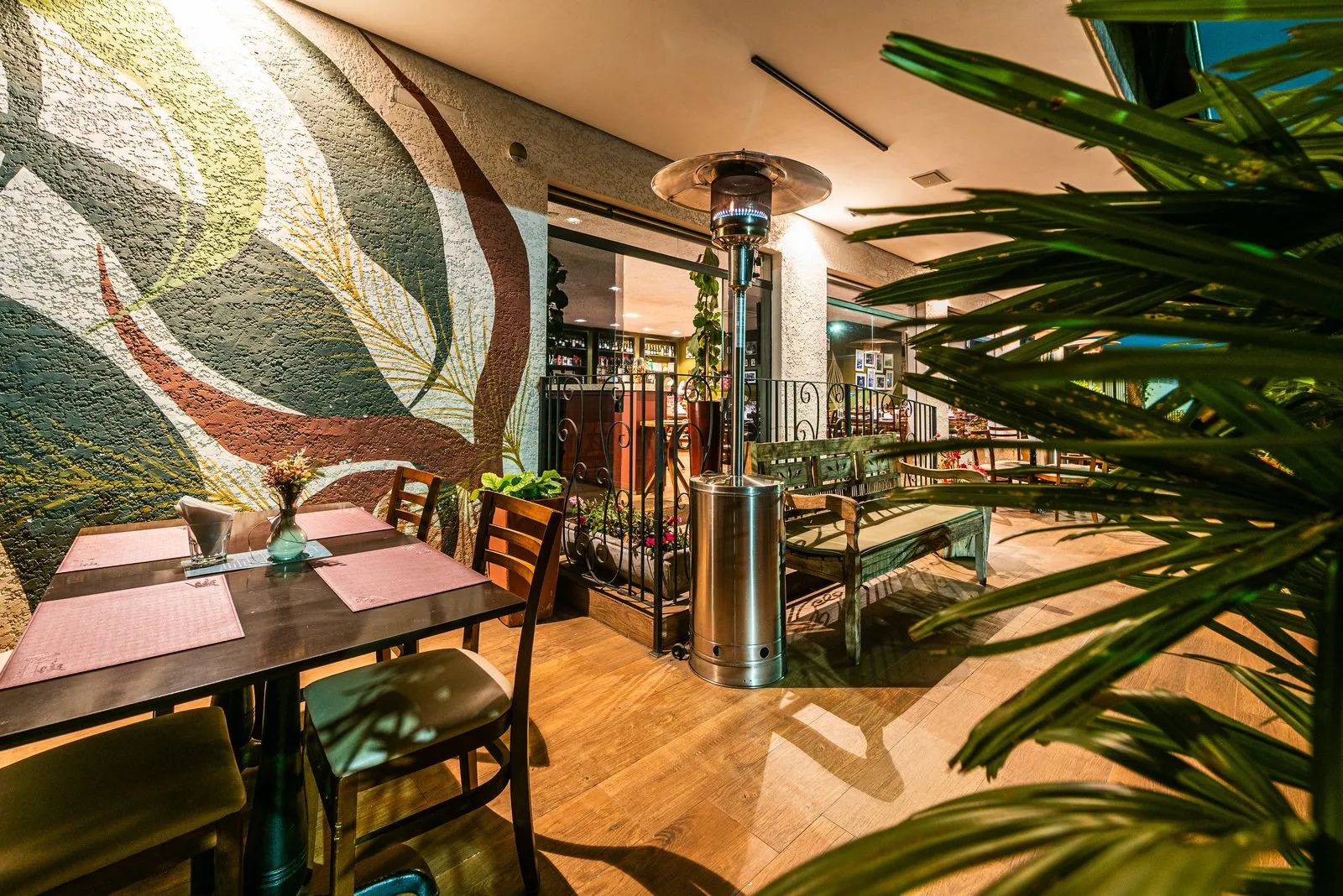 Mesa para dois na varanda da Cantina Castellammare junto a mural de folhas em verde, vinho e bege, com aquecedor aceso e grade de ferro ao fundo