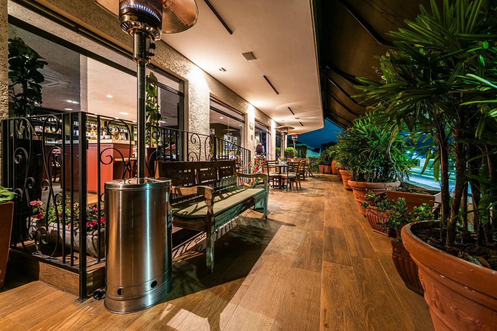 Varanda da Cantina Castellammare à noite, com banco de madeira rústica, grade de ferro, aquecedor aceso e vasos de palmeiras