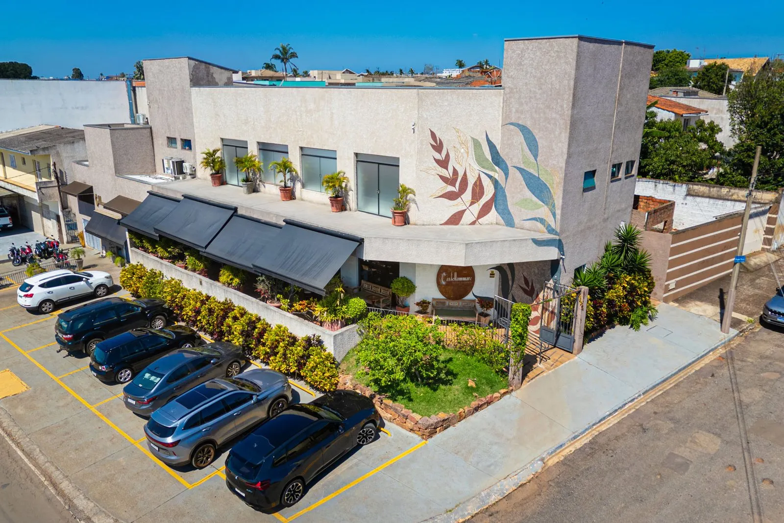 Fachada da Cantina Castellammare vista do alto, com mural de folhas, toldos pretos, varanda com plantas, jardim de esquina e carros estacionados