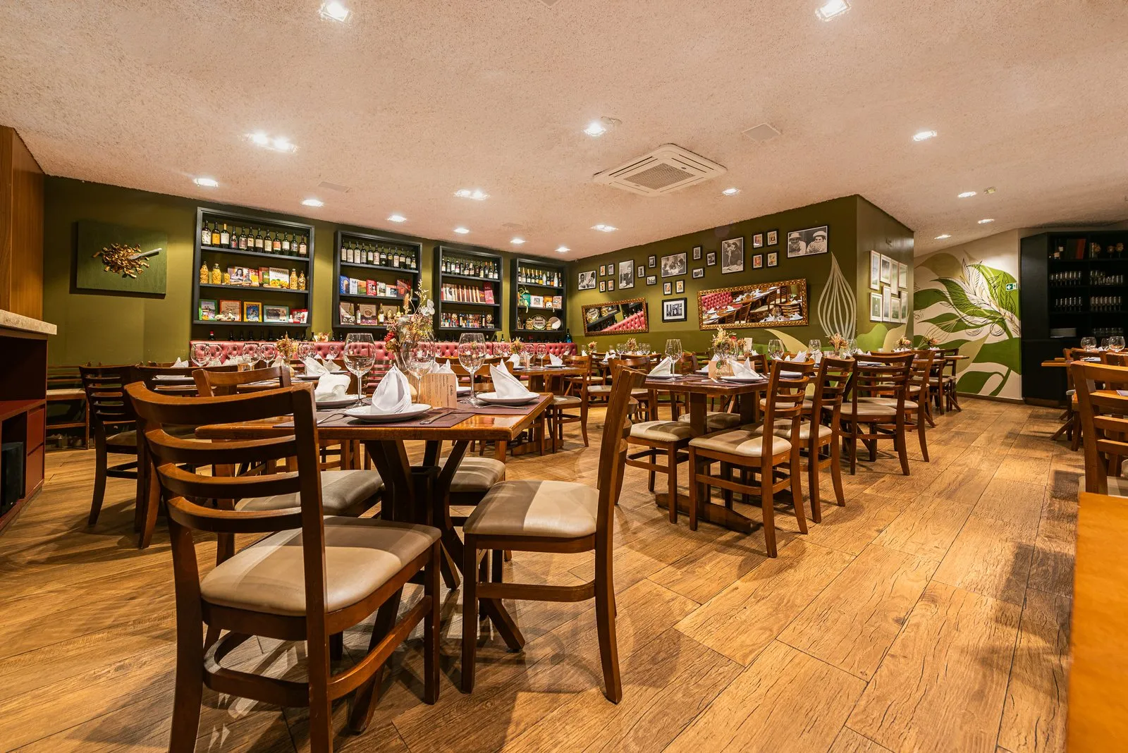 Salão da Cantina Castellammare com mesas e cadeiras de madeira, paredes verde-oliva, estantes pretas com vinhos e banco capitonê rosa ao fundo