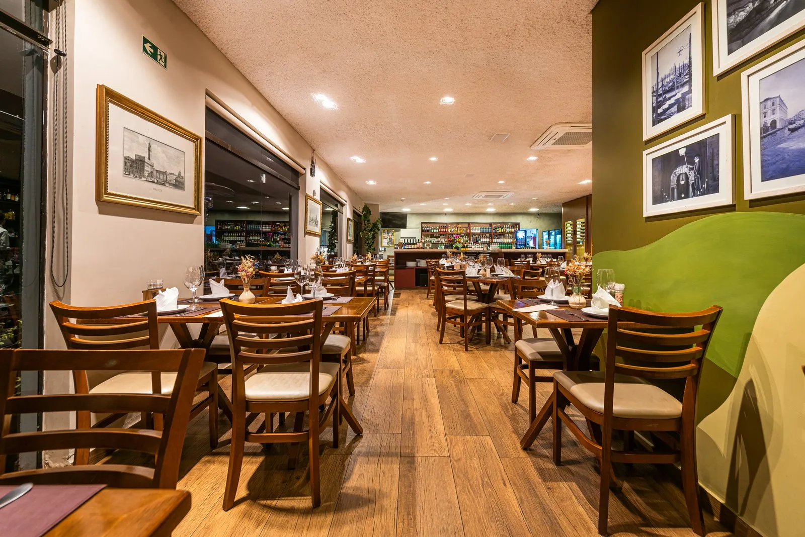 Corredor de mesas da Cantina Castellammare junto às janelas, com gravuras em moldura dourada, parede verde com fotos e o bar iluminado ao fundo
