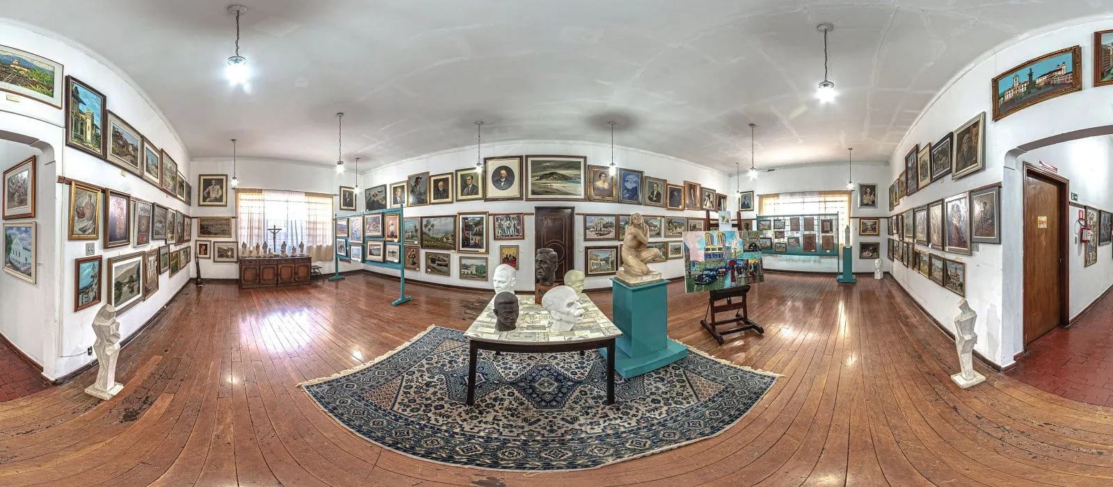 Sala do Museu Carlos Ayres com paredes cobertas de quadros, bustos sobre uma mesa central, tapete e piso de madeira