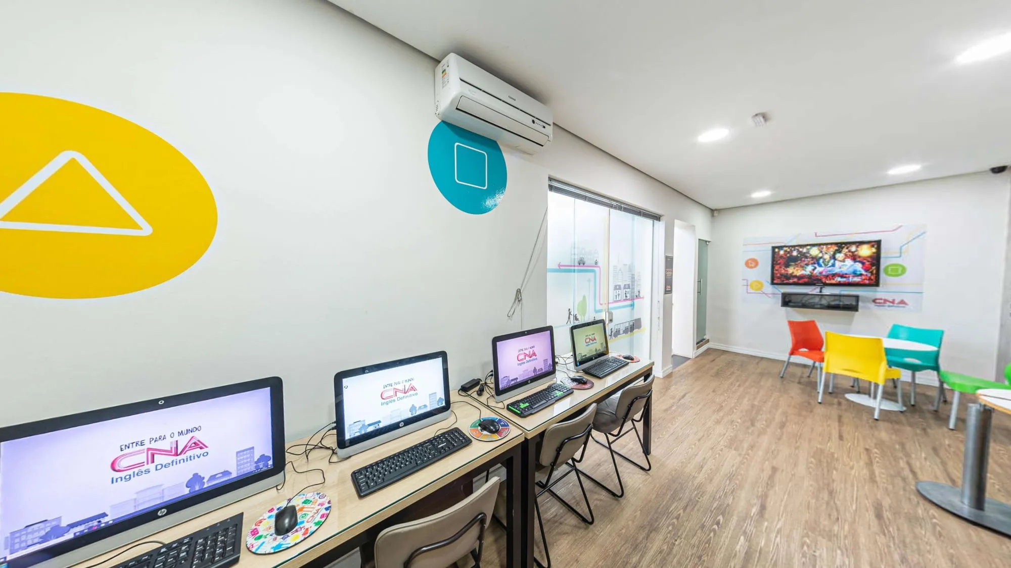 Sala de informática do CNA Itapetininga, com computadores em bancada, parede amarela e cadeiras coloridas ao fundo