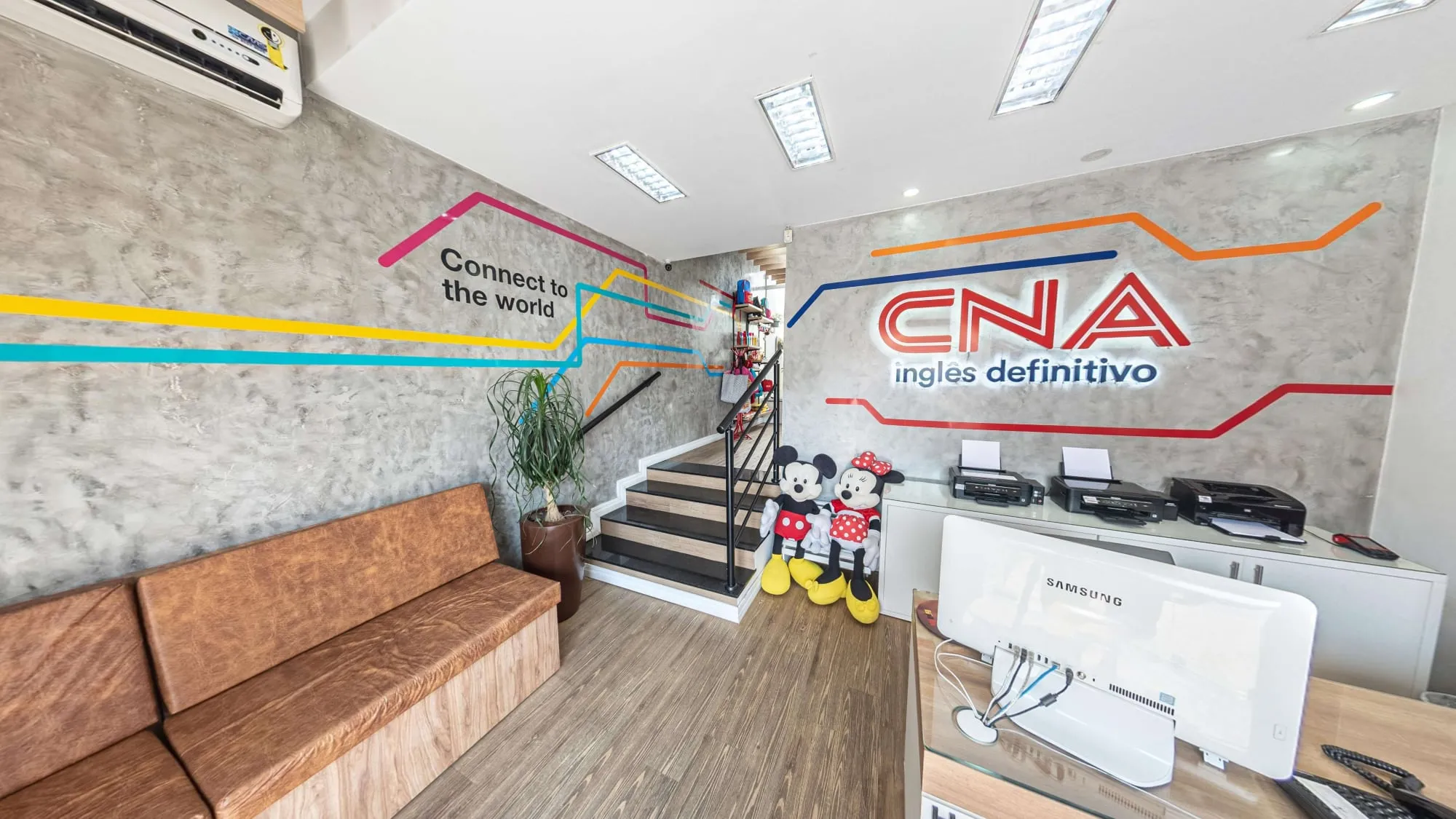 Recepção do CNA Itapetininga, com o logotipo CNA inglês definitivo na parede de cimento queimado, faixas coloridas, sofá de couro, escada e bonecos do Mickey e da Minnie junto ao balcão