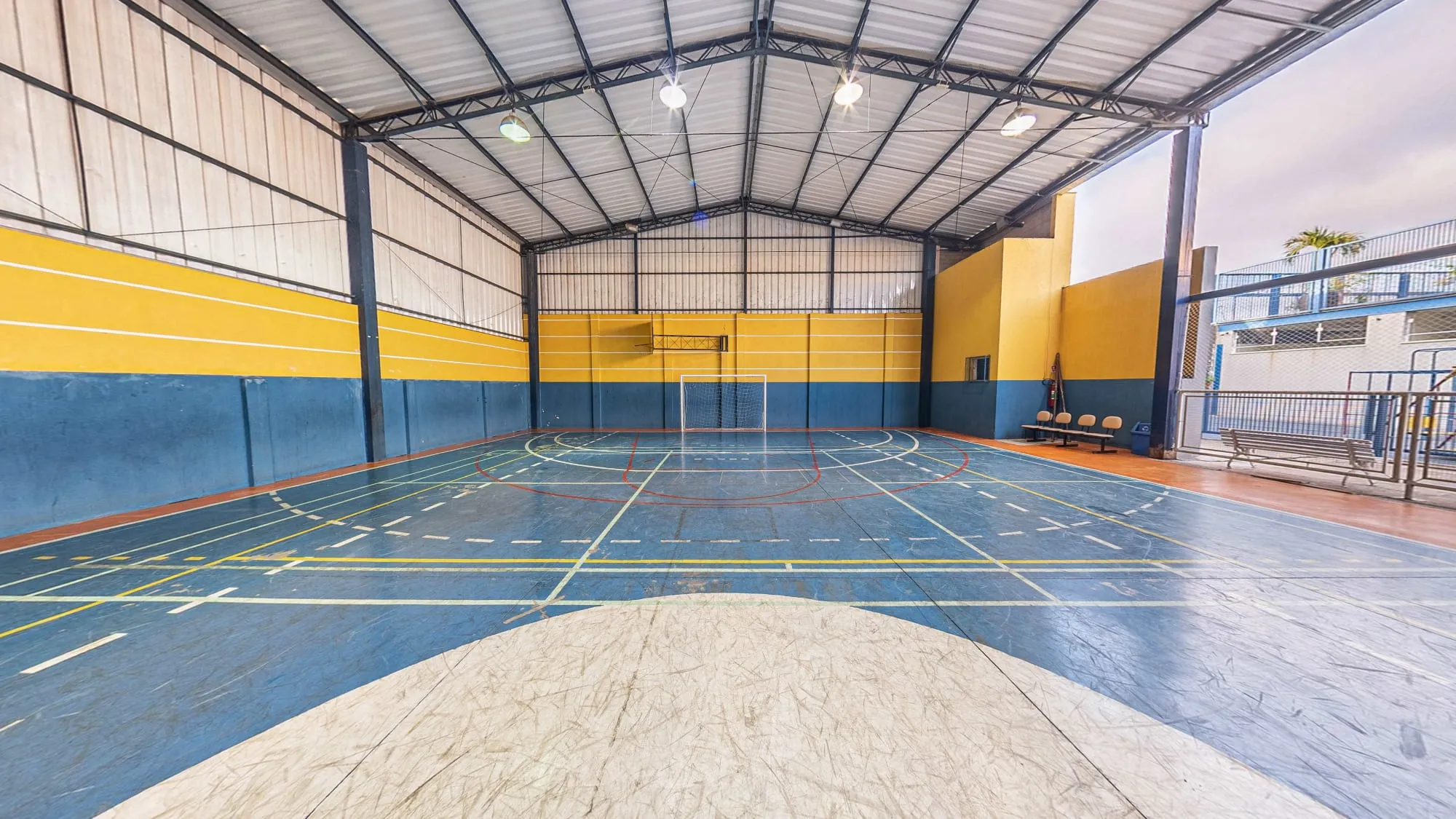 Quadra coberta do Colégio Alpis Veredas, de piso azul com marcações de várias modalidades, paredes amarelas e azuis e trave de futsal ao fundo