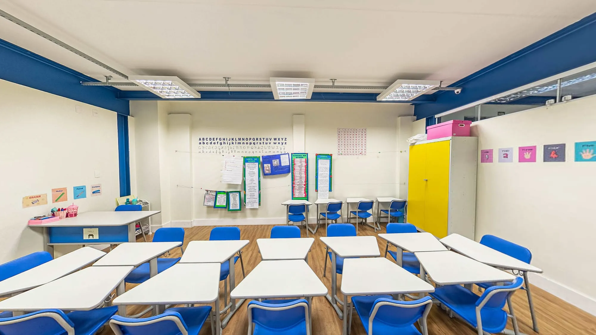 Sala infantil do Colégio Alpis Veredas, com carteiras brancas e cadeiras azuis, alfabeto e cartazes na parede e armário amarelo