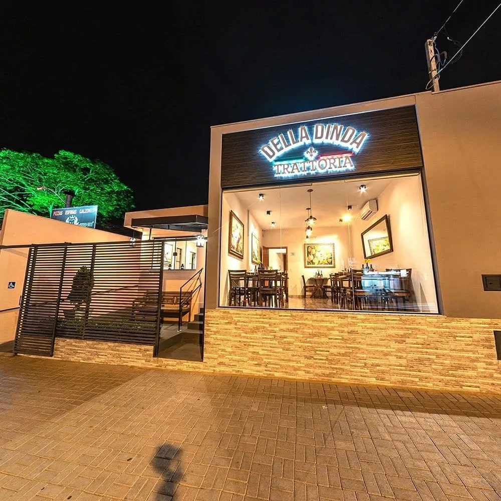 Fachada da Della Dinda Trattoria à noite, com o letreiro iluminado e o salão visível pelo vidro