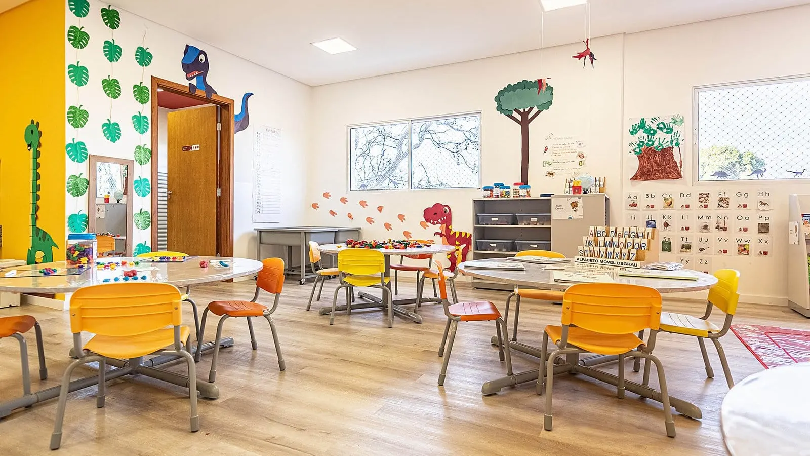 Sala de aula da educação infantil da Maple Bear Itapetininga, com mesas redondas, cadeiras amarelas e laranja, alfabeto na parede, estantes com caixas de material e dinossauros e árvores pintados nas paredes