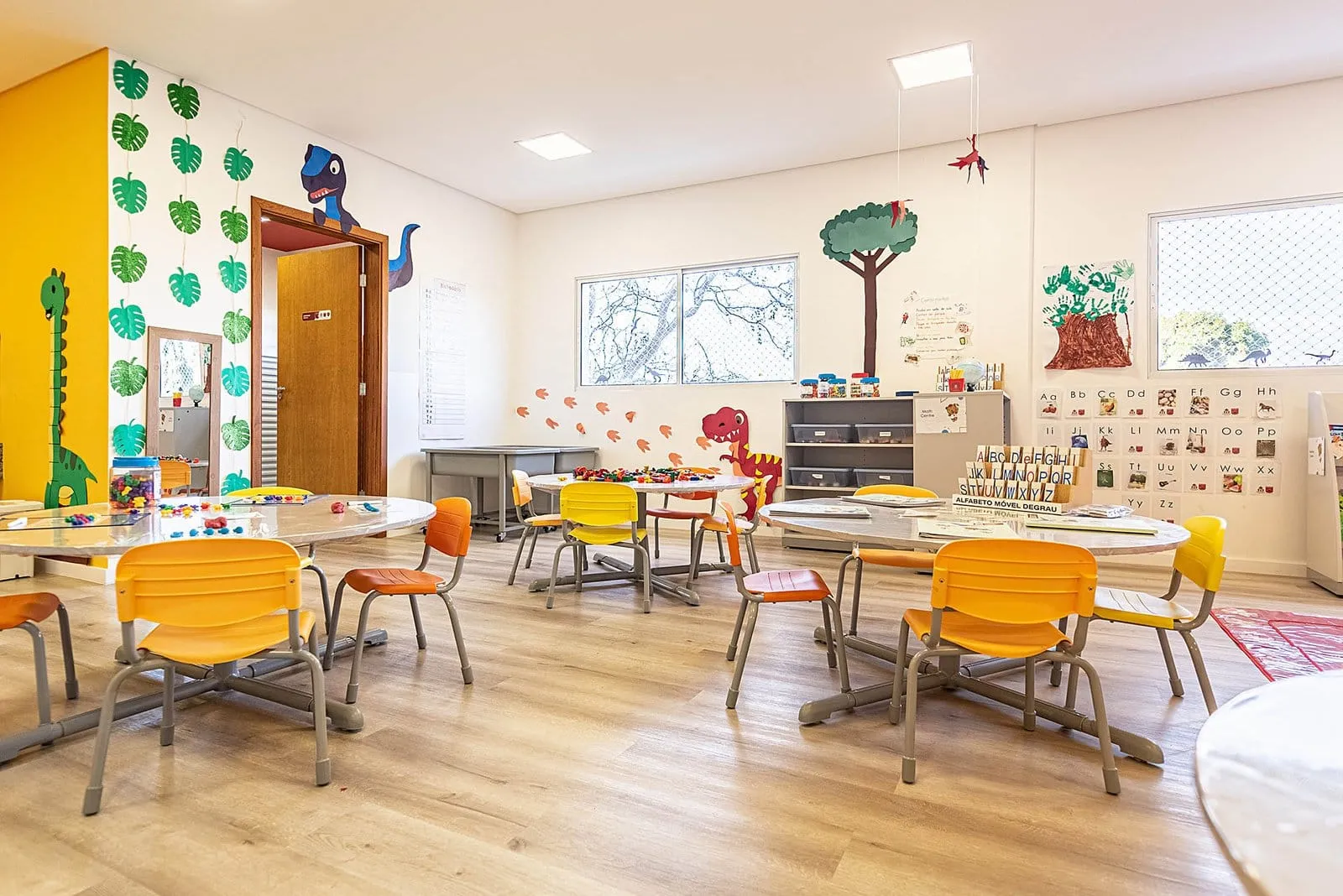 Sala de aula da Maple Bear Itapetininga com mesas redondas, cadeiras laranja e murais nas paredes