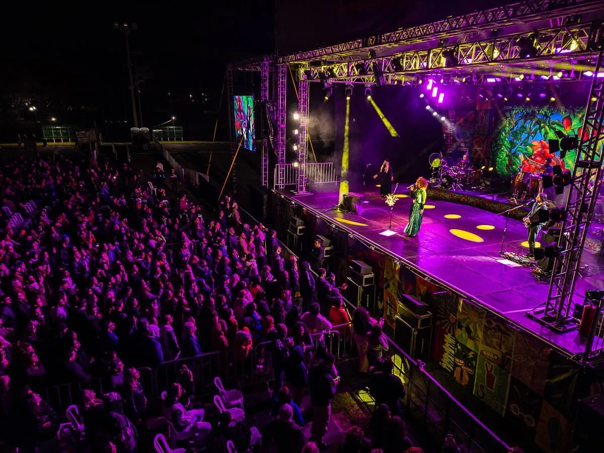 Show noturno visto do alto, com palco iluminado em roxo e a plateia ocupando o gramado