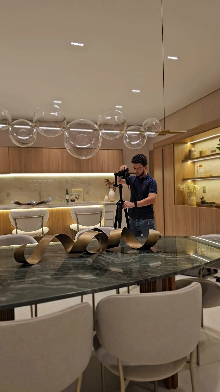Gustavo, fotógrafo da gm360, ajusta a câmera no tripé ao fundo da sala de jantar, atrás da mesa de mármore verde e do lustre de bolhas