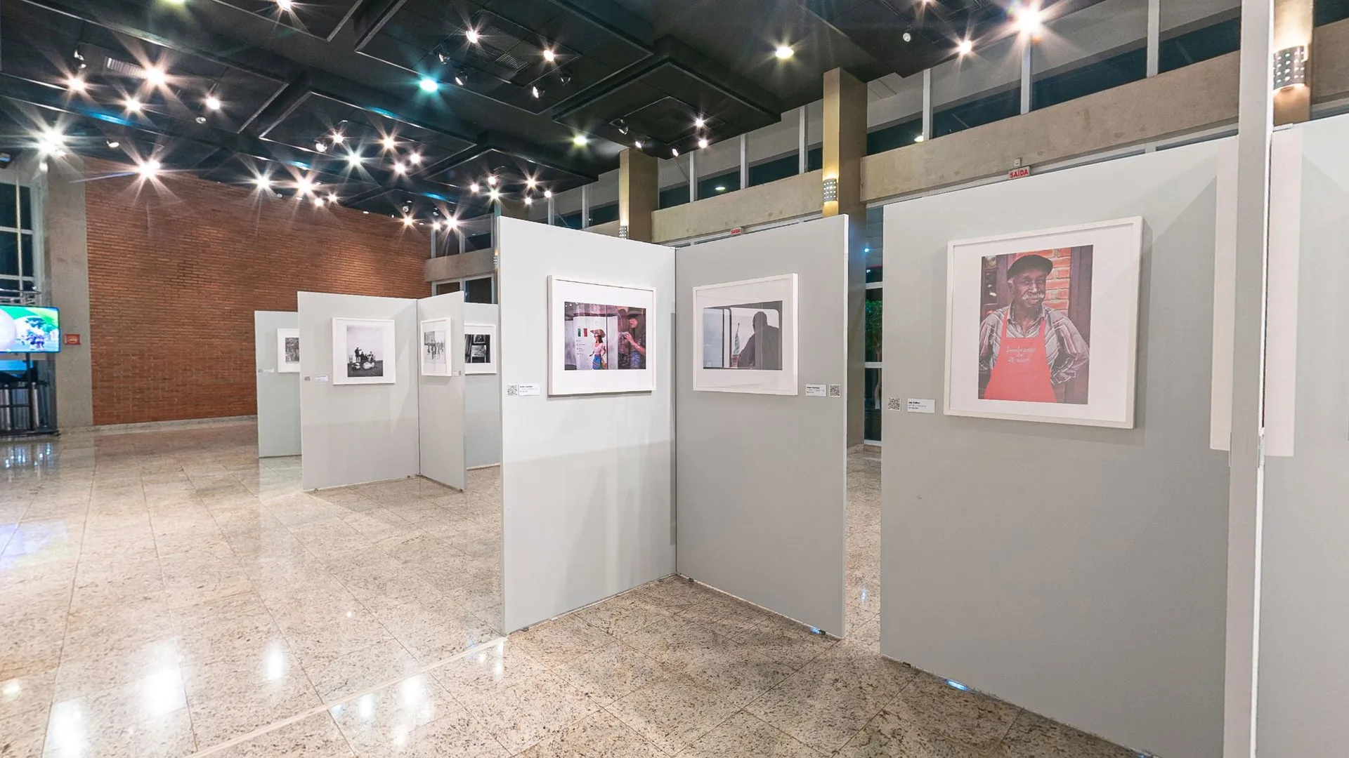 Fileira de biombos da exposição Herança Compartilhada sob o teto escuro do Foyer, com fotografias coloridas e em preto e branco