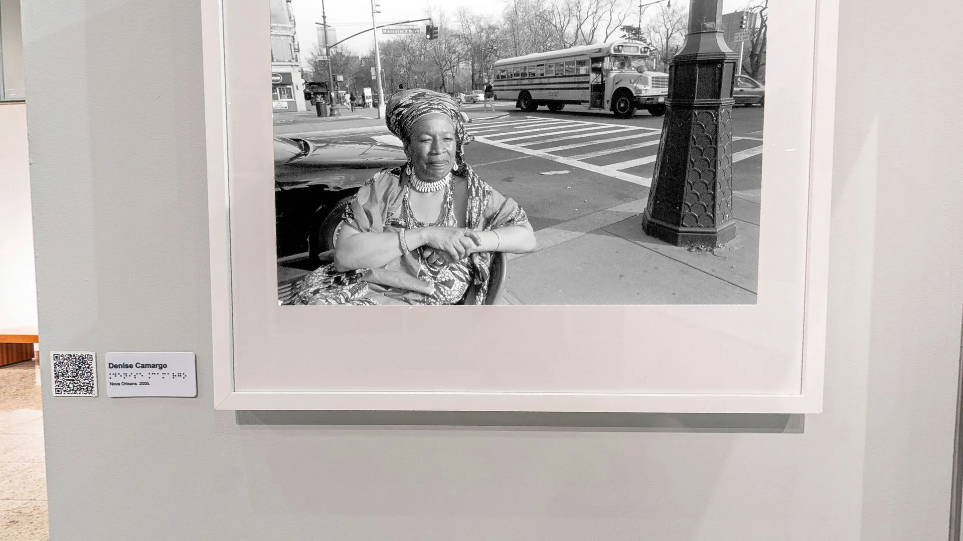 Fotografia em preto e branco de Denise Camargo com uma mulher de turbante numa esquina, emoldurada no biombo da exposição Herança Compartilhada