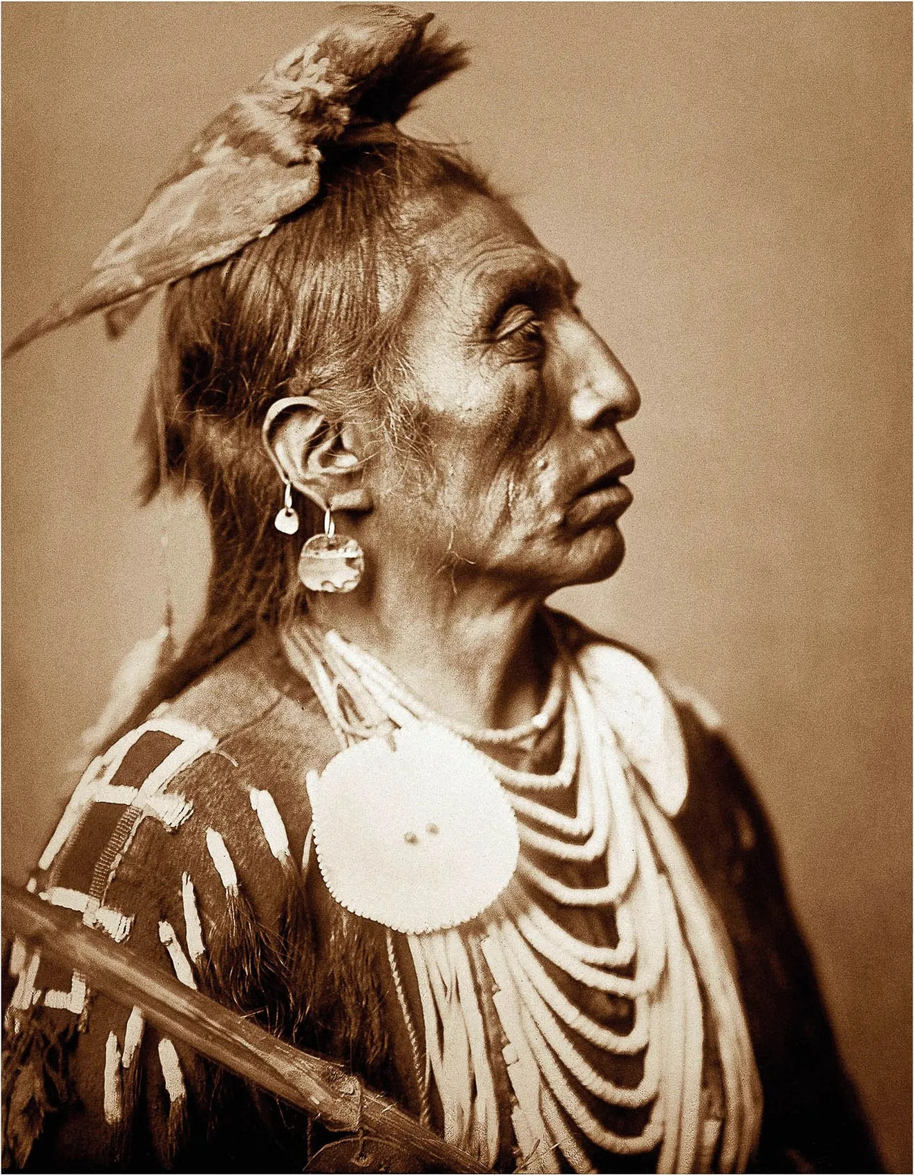 Retrato em sépia de um pajé Crow de perfil, com colares de conchas em camadas, um grande disco branco no peito e uma pele de animal amarrada no alto da cabeça, sobre fundo neutro