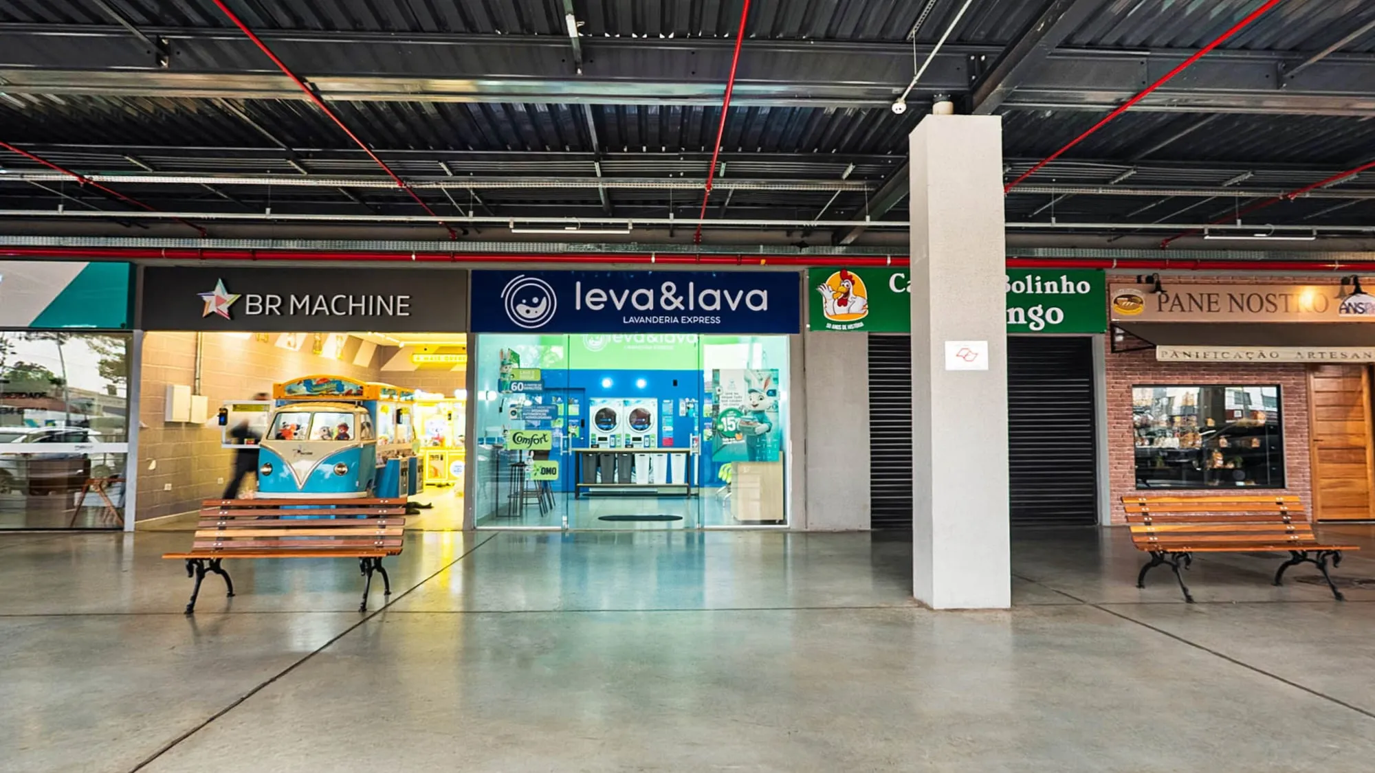 Fachada da Leva & Lava na galeria do Tenda Atacado, com letreiro azul-marinho, vitrine de vidro mostrando as máquinas ao fundo e bancos de madeira na calçada coberta