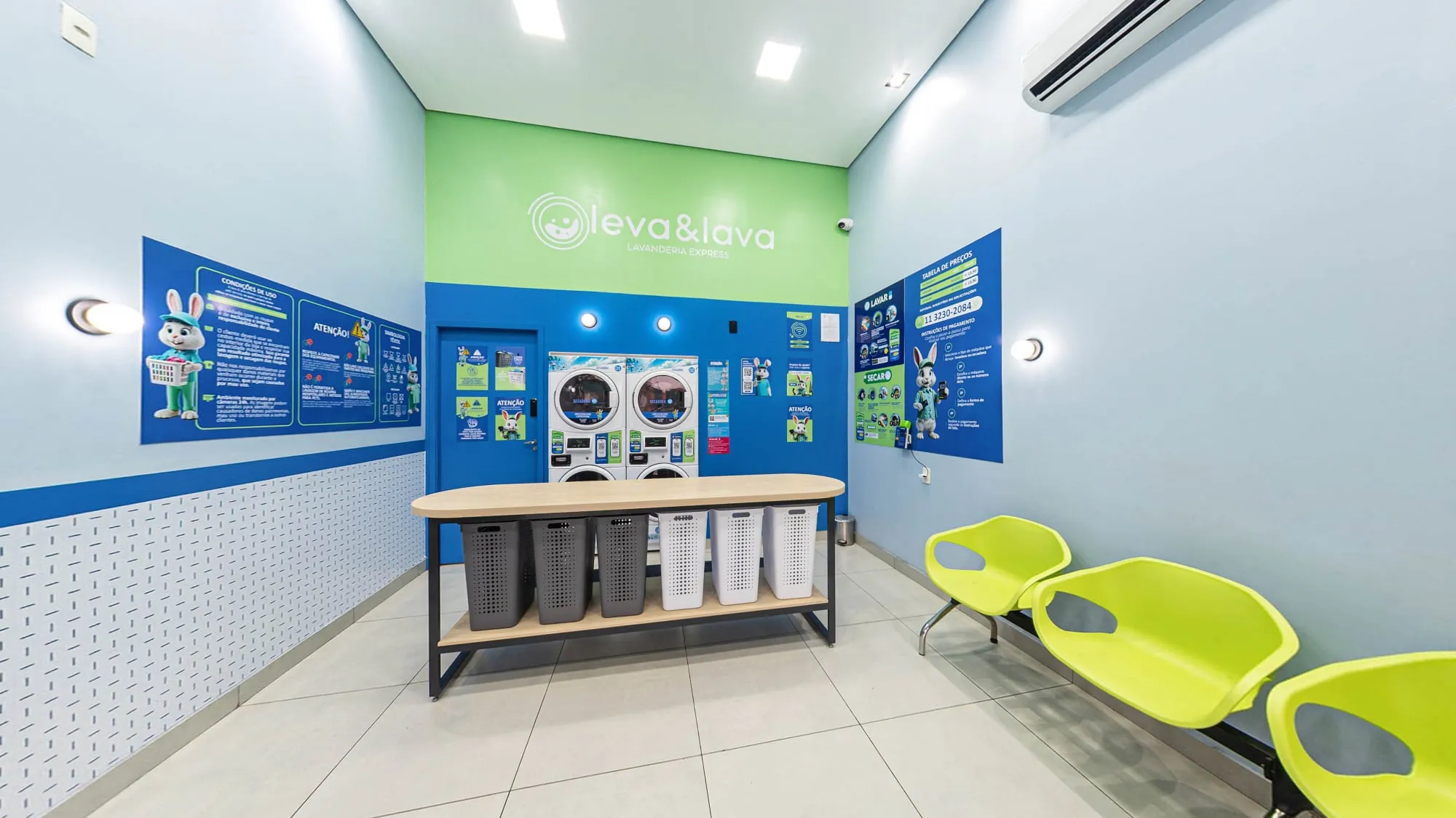 Interior da Leva & Lava em Itapetininga, com paredes azul-claras, a faixa verde com o logotipo ao fundo, as lavadoras e secadoras empilhadas, a bancada de cestos cinza e brancos e cadeiras verde-limão