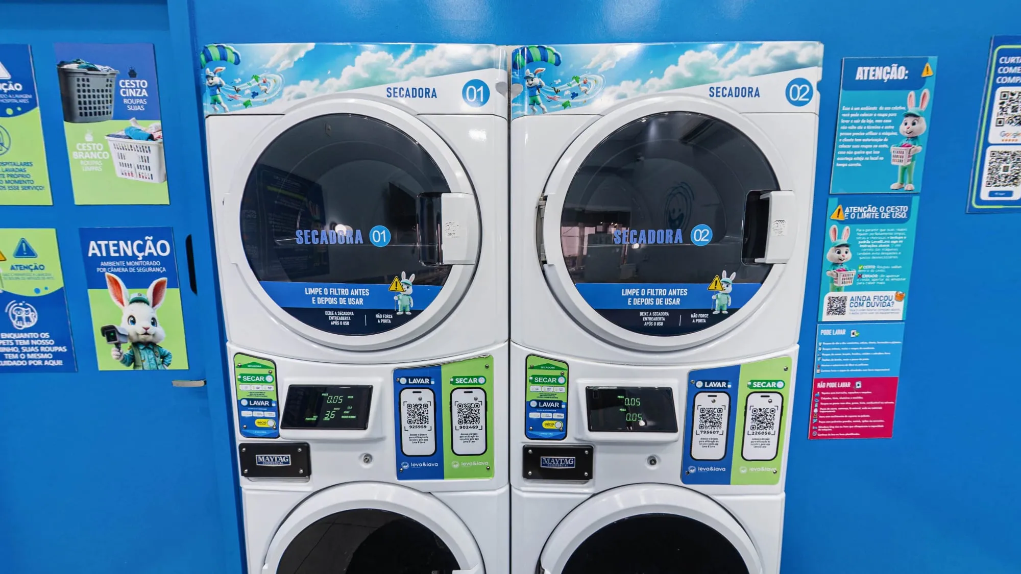 Duas secadoras empilhadas sobre lavadoras, com adesivos azuis numerados, QR Codes de instrução no painel e cartazes de aviso com o coelho mascote ao lado
