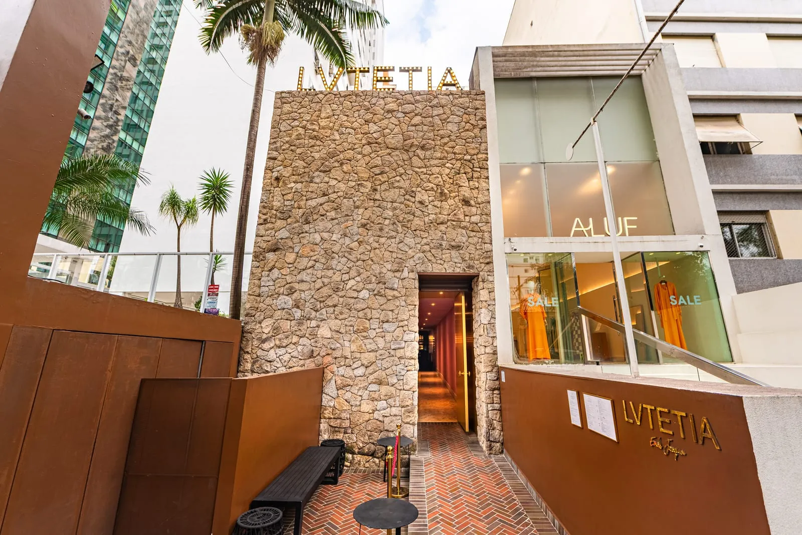 Entrada do Lvtetia na rua, com parede de pedra, letreiro dourado iluminado no alto, corredor rosado e piso de tijolos em espinha de peixe