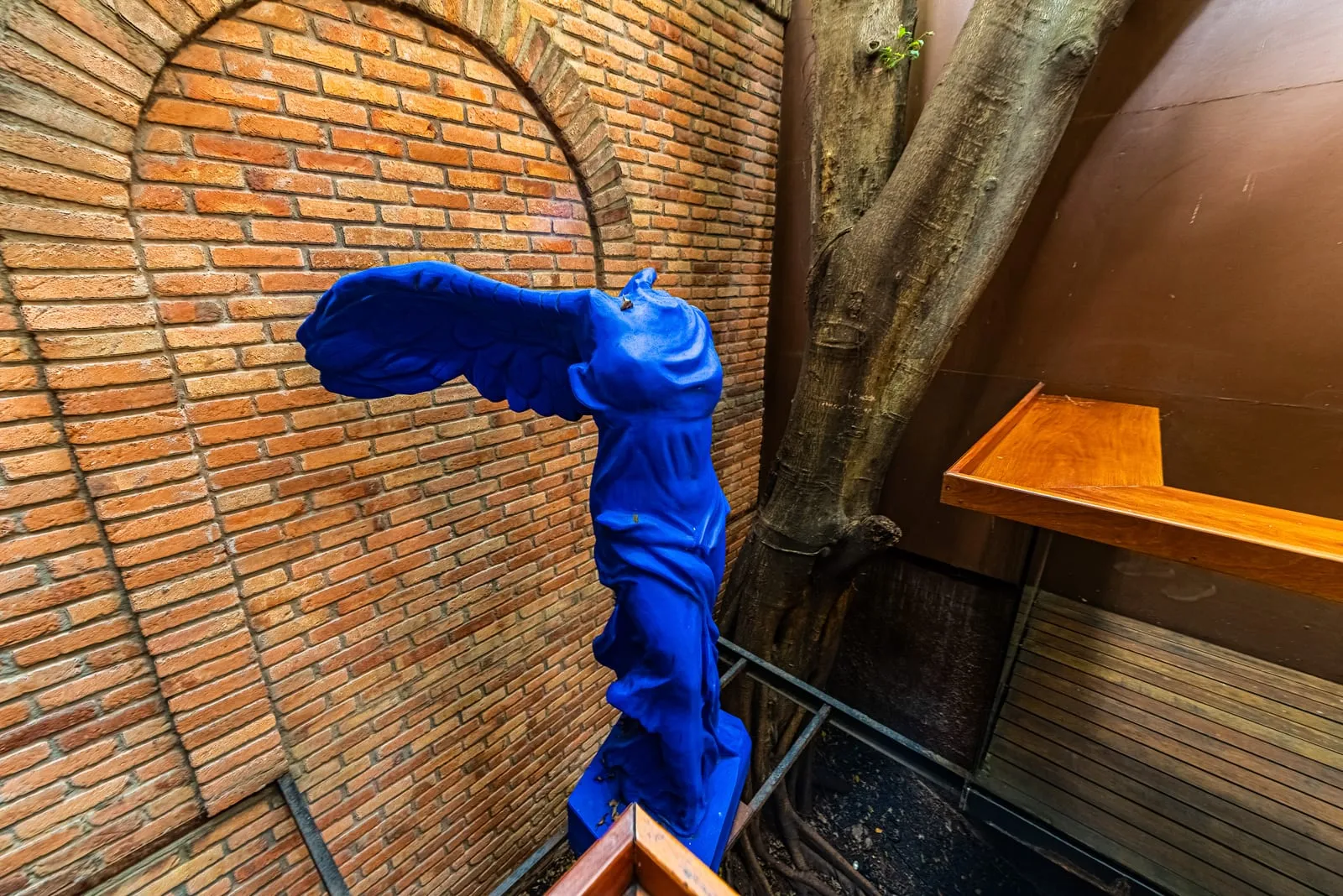 Réplica azul da Vitória de Samotrácia diante de parede de tijolos com arco, ao lado do tronco de uma árvore
