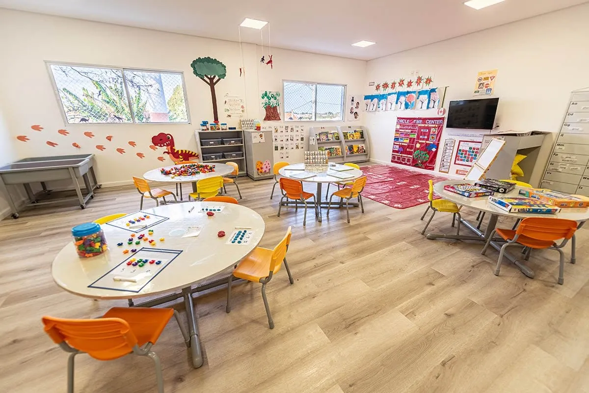 Sala com mesas redondas, cadeiras laranja, materiais de pintura e estantes com livros