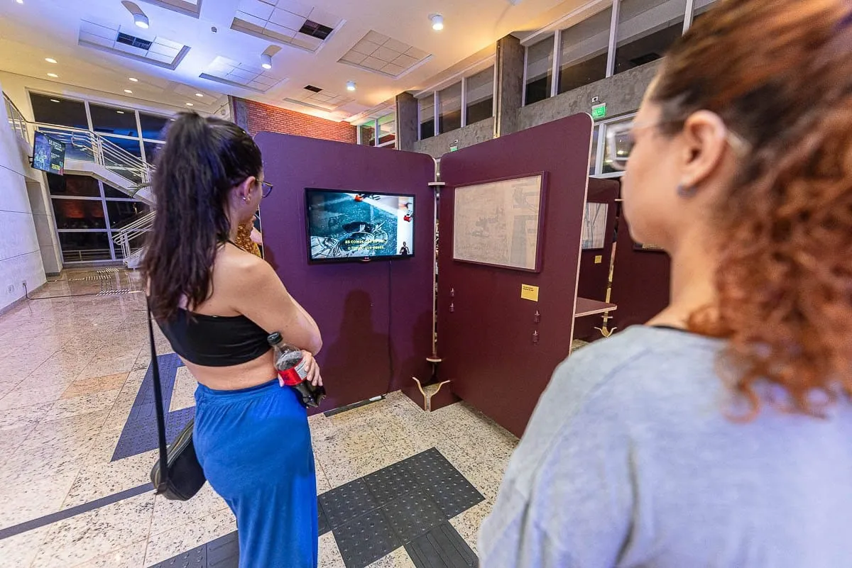 Visitantes assistem a vídeo em monitor entre os painéis da exposição Terra Terreno Território