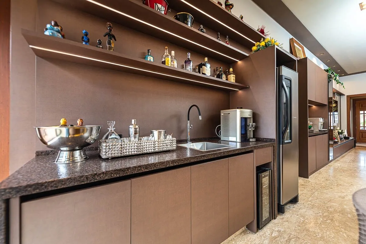 Bar com bancada escura, prateleiras com garrafas e cuba de inox, integrado à cozinha