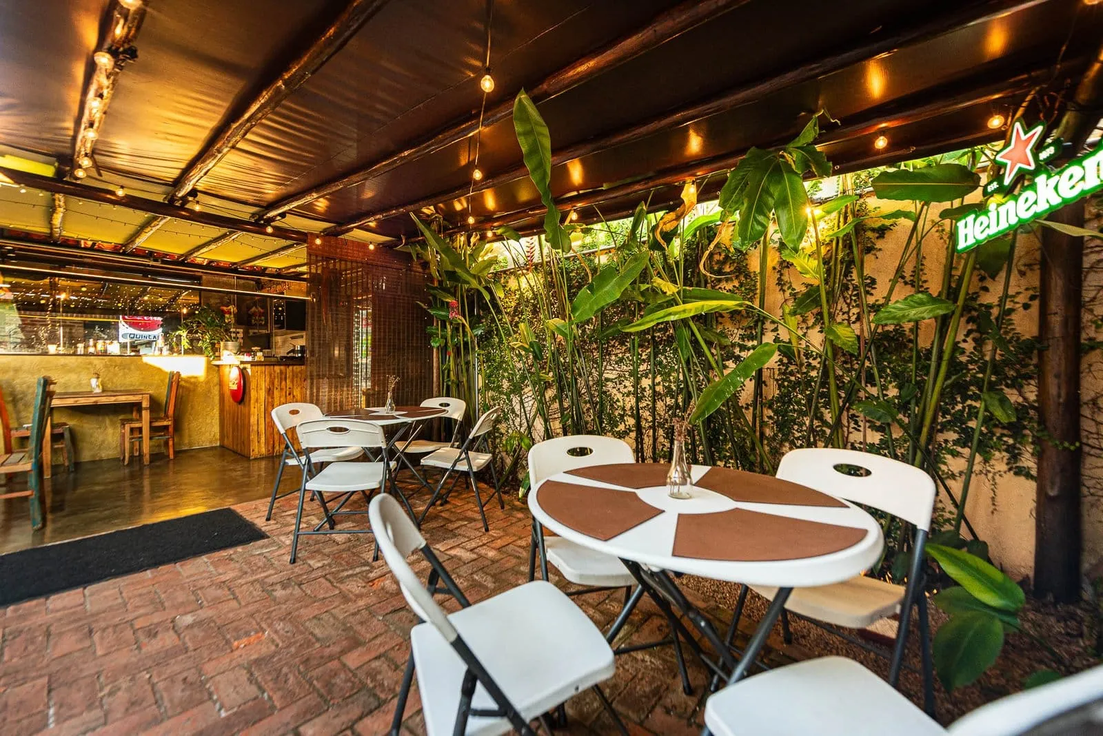 Área externa do Quintal Iwasa'i com mesas e cadeiras brancas, plantas tropicais e balcão de bar ao fundo