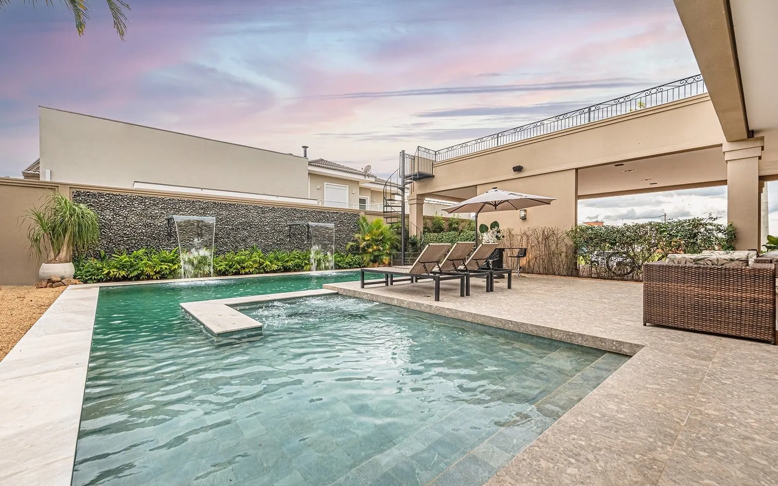 Piscina da Residência R&M ao entardecer, sob céu rosado, com cascatas no muro de pedra, espreguiçadeiras e varanda coberta