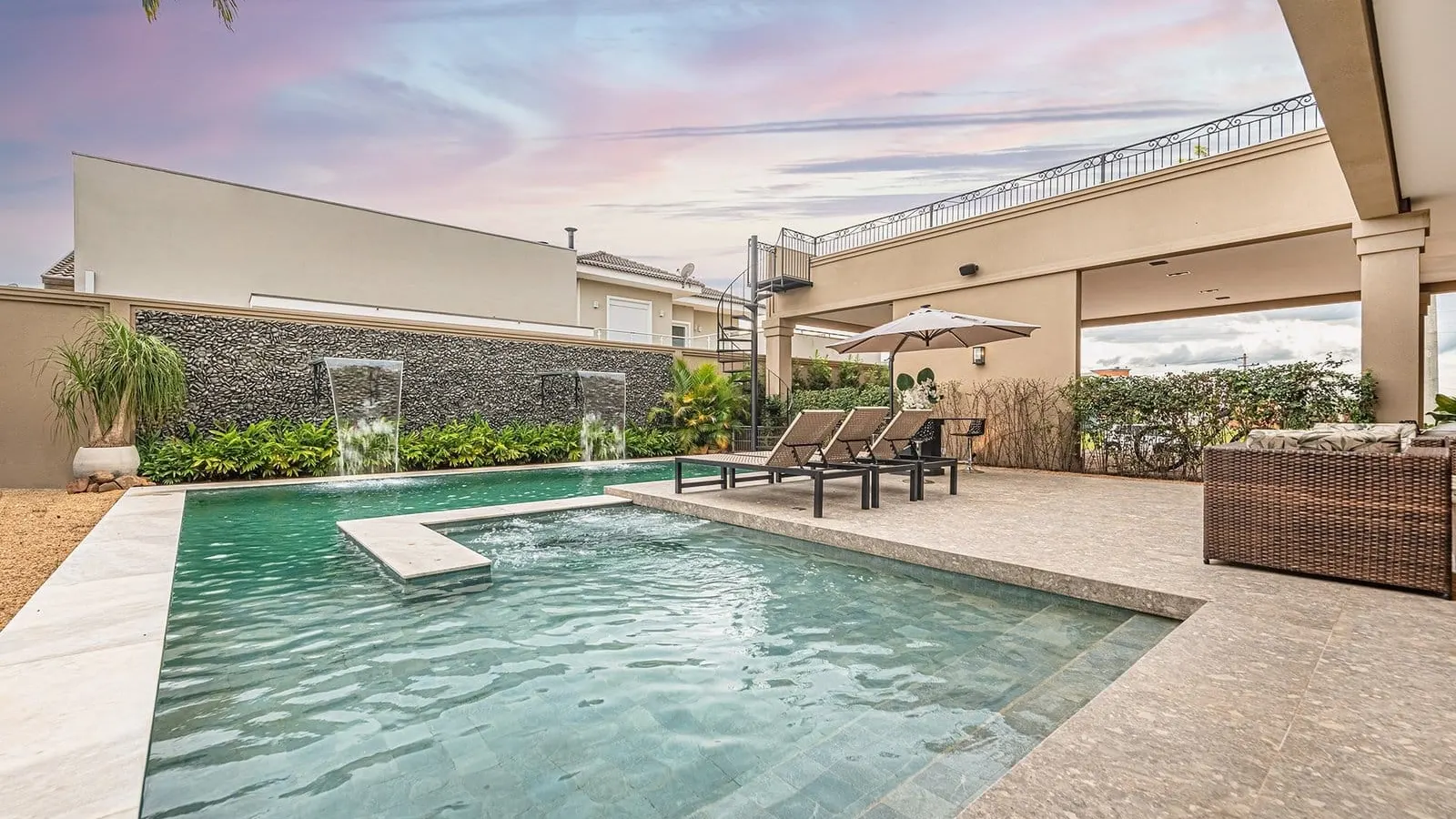Piscina da Residência R&M ao entardecer, sob céu rosado, com cascatas no muro de pedra, espreguiçadeiras e varanda coberta