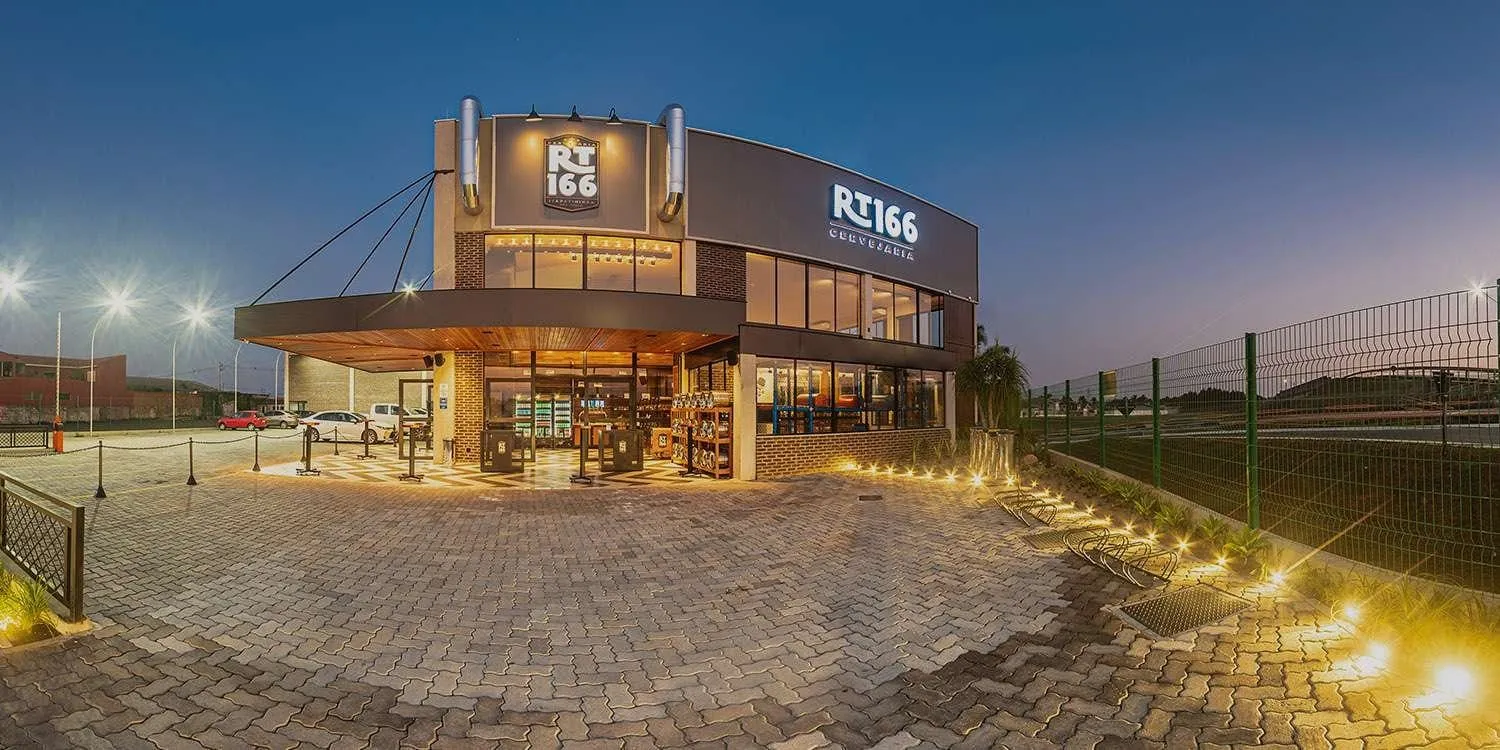Fachada da RT166 Cervejaria ao anoitecer, com letreiro iluminado, entrada envidraçada e pátio de pedra