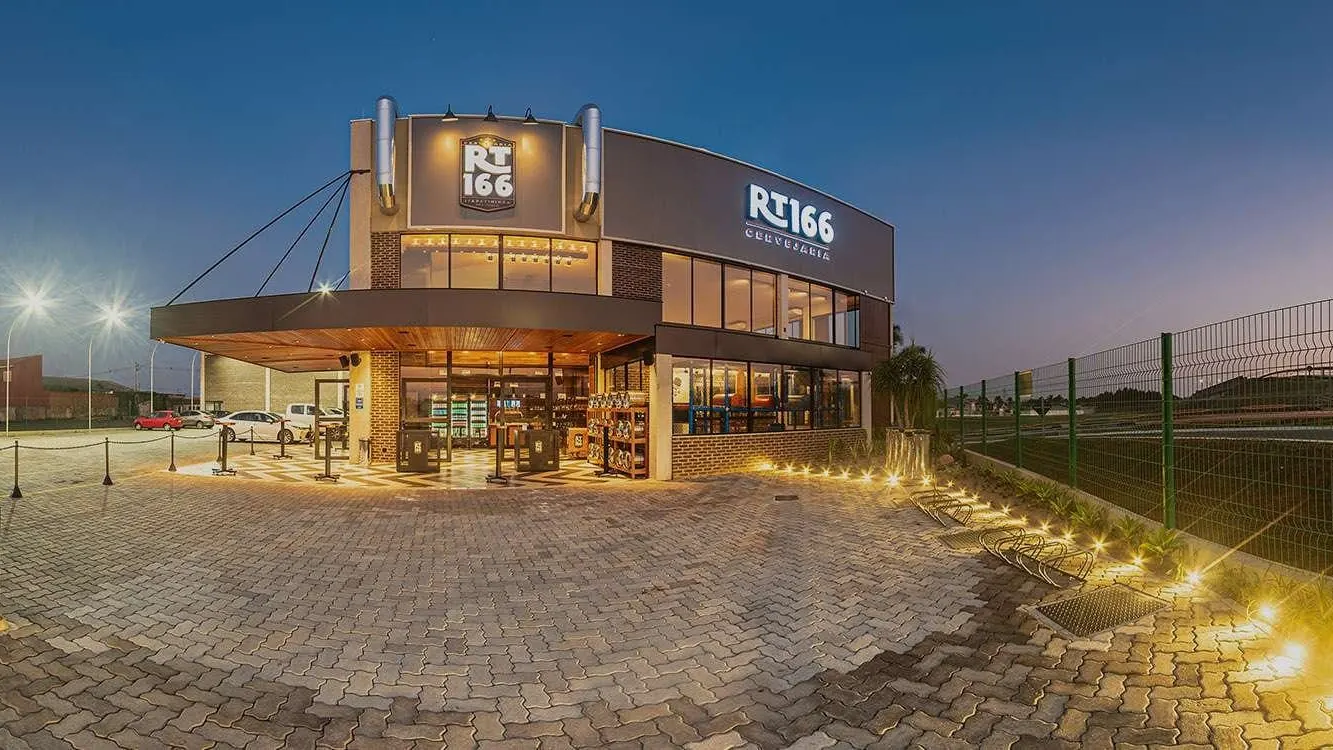 Fachada da RT166 Cervejaria, em Itapetininga, ao anoitecer, com letreiro iluminado, entrada envidraçada e pátio de pedra