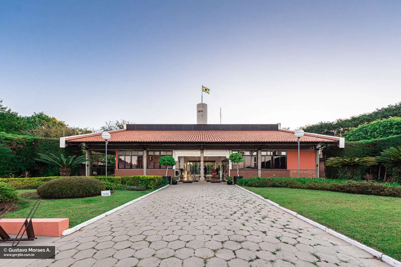 Fachada do SESI Itapetininga com caminho de pedra, jardim e torre com a bandeira do Brasil