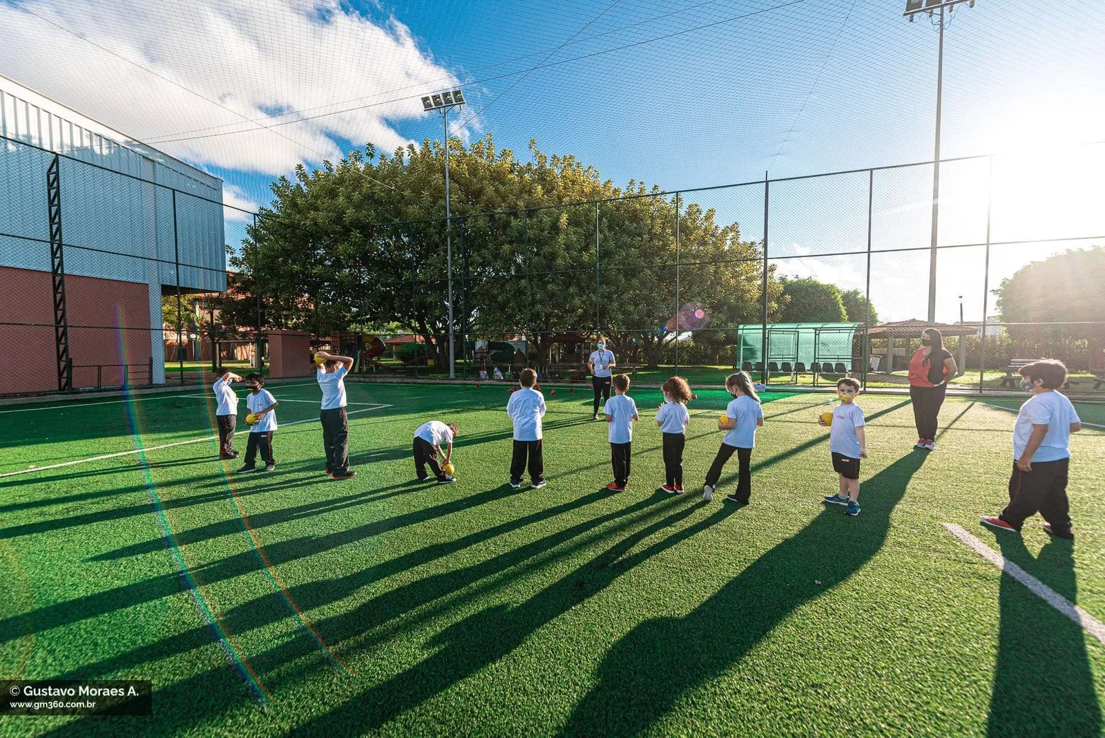 Alunos em atividade no campo de grama sintética, com sombras longas no fim da tarde