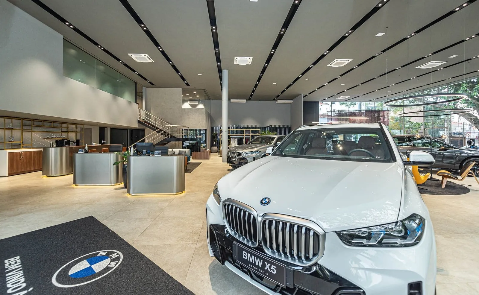Salão da BMW Osten Perdizes com um BMW X5 branco em primeiro plano, balcões de atendimento e escada para o mezanino ao fundo