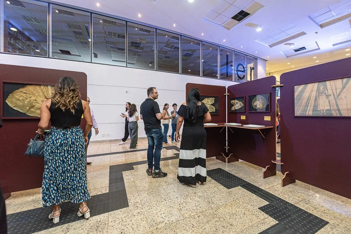 Visitantes circulam entre os painéis com impressões em folhas e fotografias, sobre piso com faixa tátil